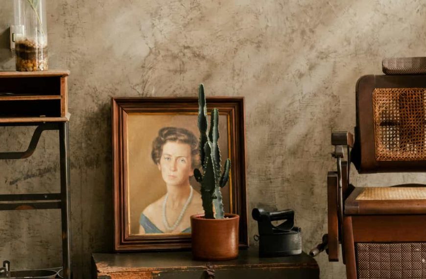 Framed portrait of a woman with a pearl necklace, leaning against a textured wall, cactus plant in a clay pot in front, antique wooden chair with cane backing, and old furniture surrounding the scene