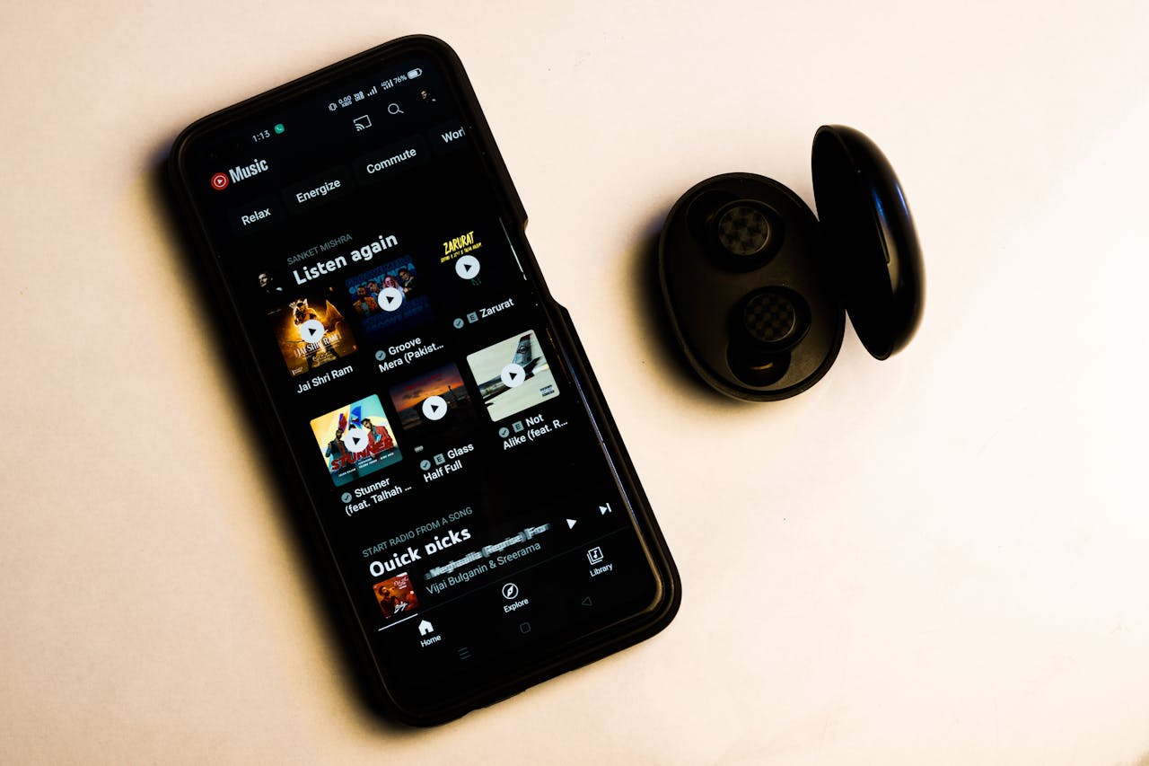 Smartphone displaying a music streaming app, black wireless earbuds in open charging case beside the phone, white background, screen shows album covers and playlists