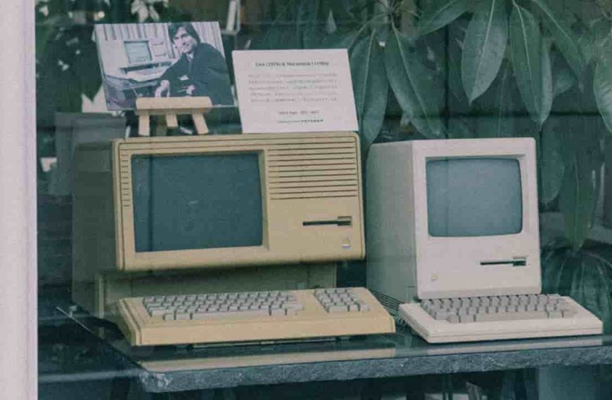 Vintage Apple Lisa and Macintosh computers, beige casing, old-style CRT screens, physical keyboards, displayed on a dark table behind glass, with a photo of Steve Jobs and an information card