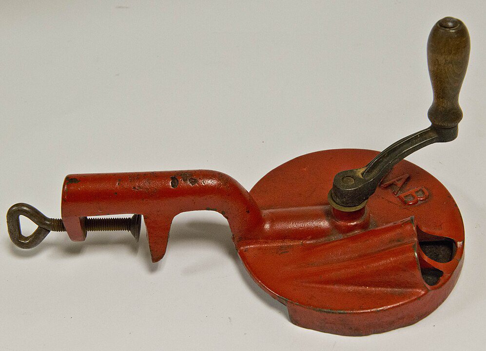 Vintage red hand-crank apple peeler, metal body with rotating handle, clamp base for table attachment, spiral blade slot, used for peeling and coring apples