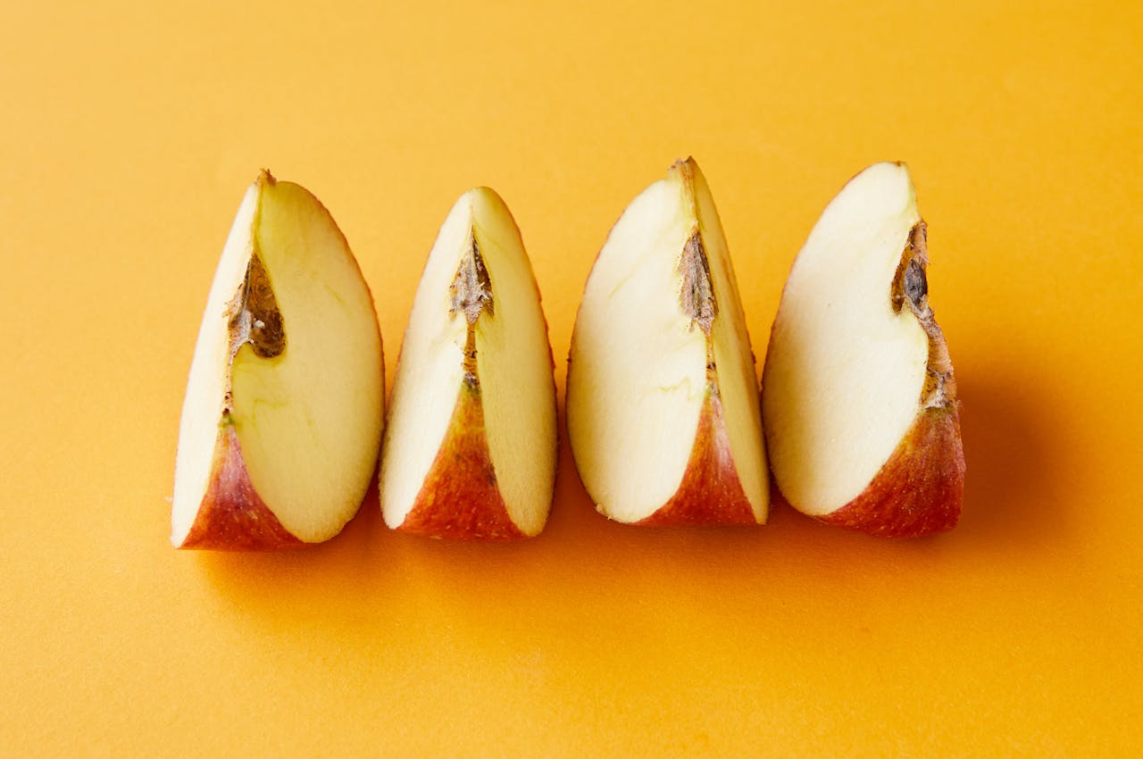 Four red apple slices arranged in a row on a bright yellow background, showing white flesh and brown seeds. Each wedge displays the curved outer red skin and clean-cut edges