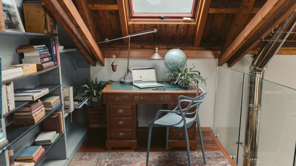 Cozy attic study with sloped wooden ceiling, skylight window, wooden desk with green inlay, open laptop, desk lamp, globe, potted plants, modern gray chair, bookshelf filled with books, and a patterned rug on the floor