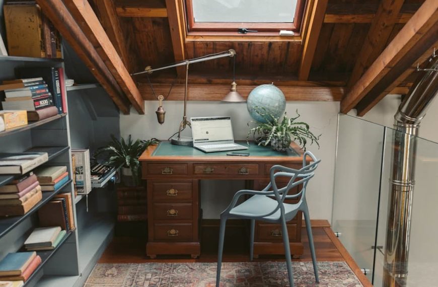 Cozy attic study with sloped wooden ceiling, skylight window, wooden desk with green inlay, open laptop, desk lamp, globe, potted plants, modern gray chair, bookshelf filled with books, and a patterned rug on the floor