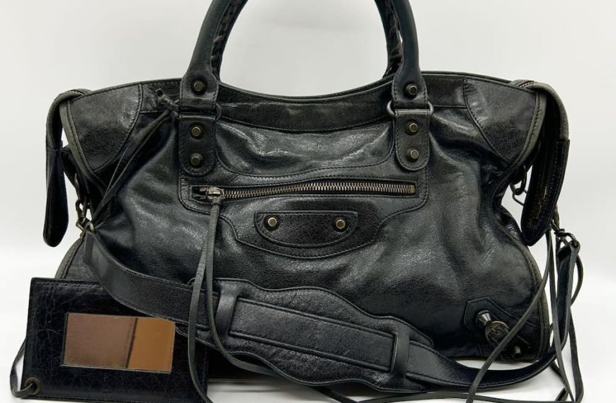 A well-worn black leather handbag with studded details, zippered pockets, and dual handles sits on a white surface. A small black wallet with brown leather interior is positioned beside the bag.