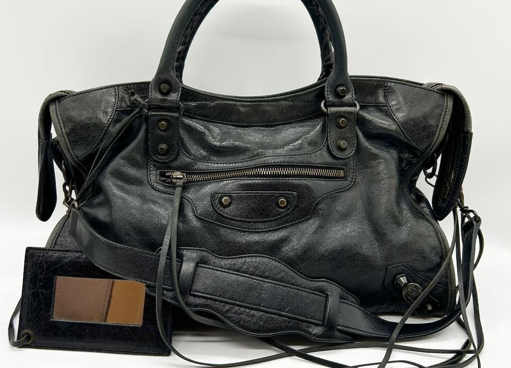 A well-worn black leather handbag with studded details, zippered pockets, and dual handles sits on a white surface. A small black wallet with brown leather interior is positioned beside the bag.