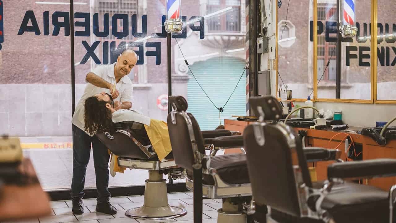 Barber in a white shirt giving a shave to a client reclining in a chair at a classic-style barbershop, with empty chairs and tools visible around the shop