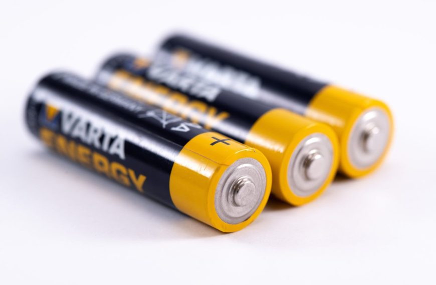 Three AA batteries, black and yellow casing, arranged in a row, silver terminals facing forward, white background