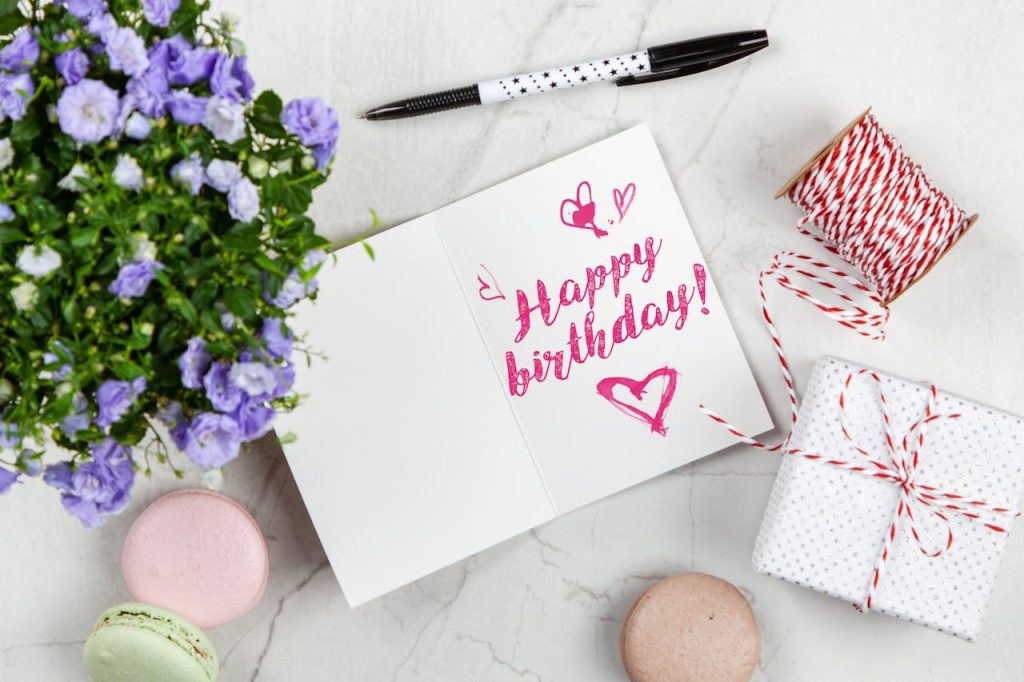 Birthday card with “Happy Birthday!” written in pink, open on a marble surface, surrounded by a pen, purple flowers in a pot, string, a wrapped gift