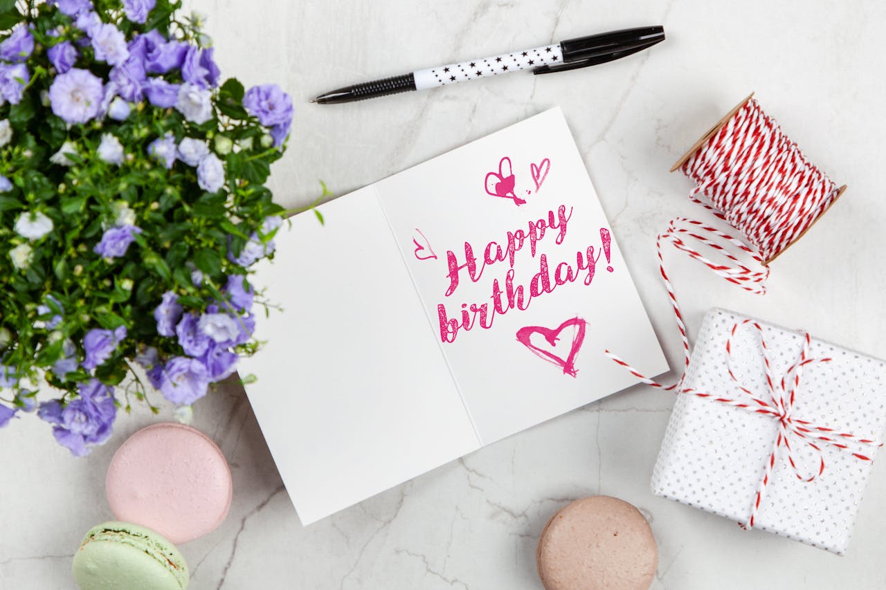 Birthday card with “Happy Birthday!” written in pink, open on a marble surface, surrounded by a pen, purple flowers in a pot, string, a wrapped gift