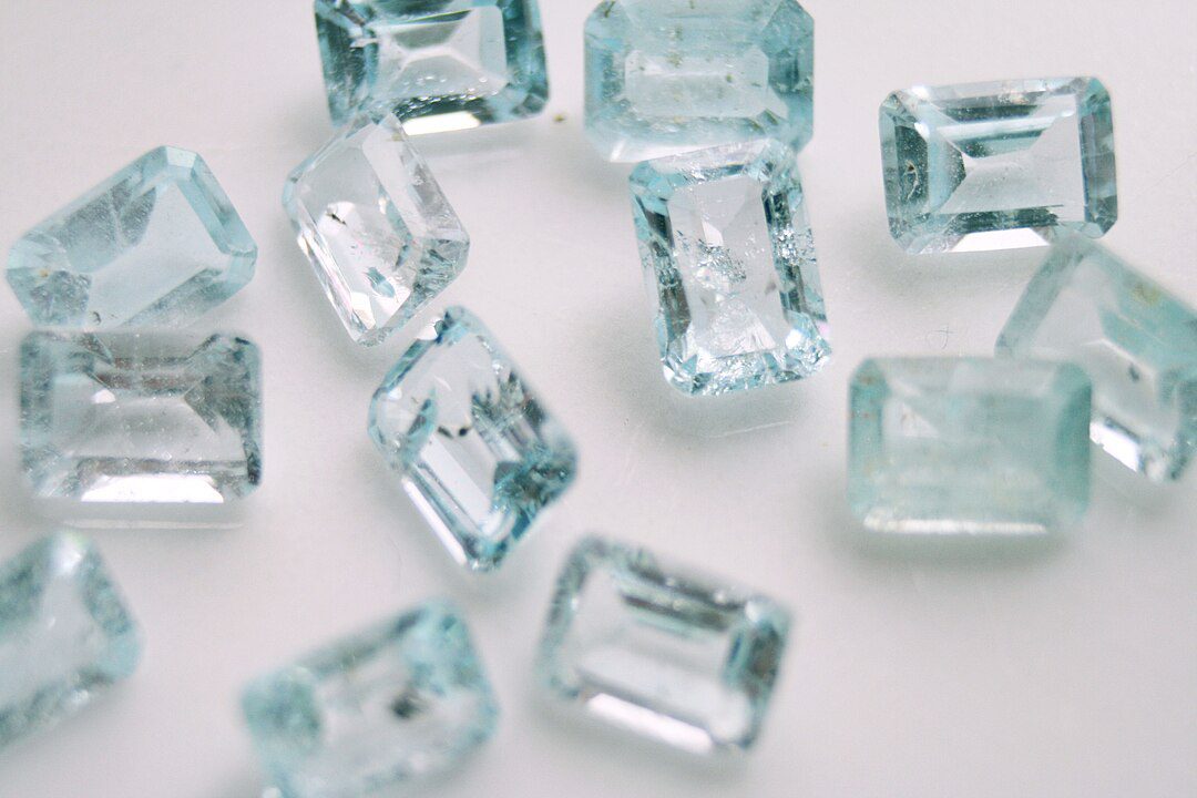 Scattered aquamarine gemstones, rectangular cuts with light blue hue, placed on a smooth white surface, transparent and reflective, showcasing brilliance and clarity