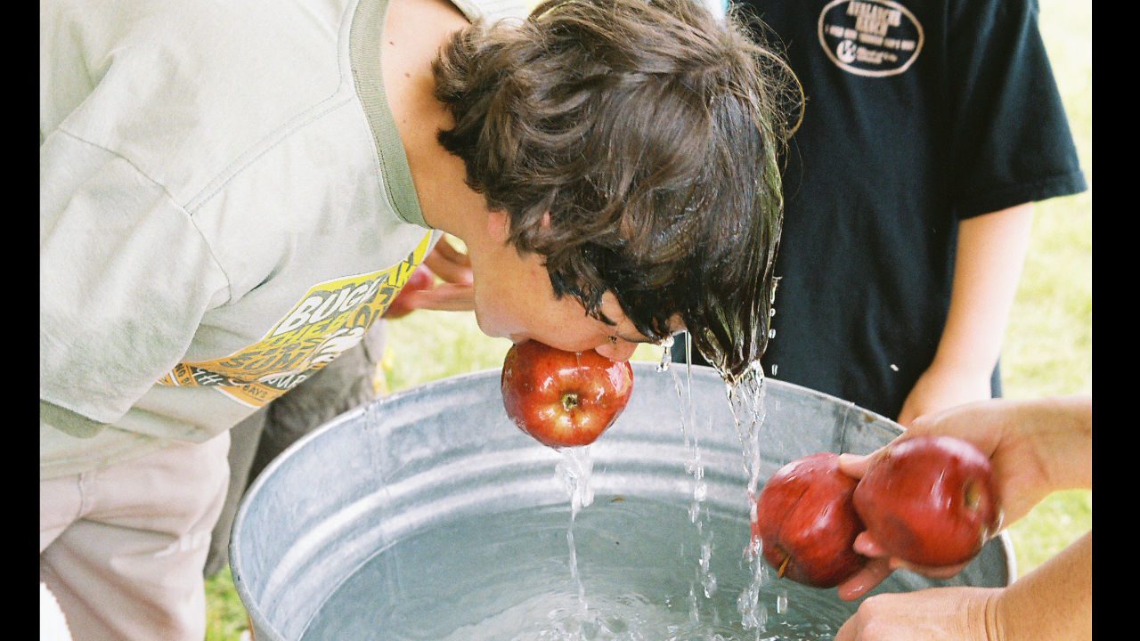 Boy bobbing for apples in a metal tub filled with water, surrounded by others holding red apples