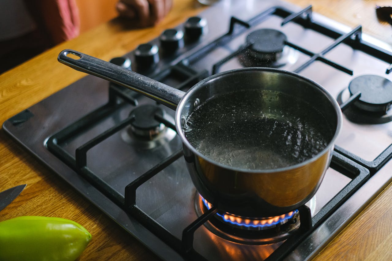 Stainless steel pot on a lit gas stove, boiling clear liquid, kitchen countertop with a green bell pepper and knife nearby, visible burner flames