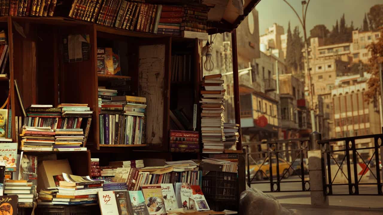 Outdoor bookstall filled with stacks of books in Arabic, with wooden shelves and a backdrop of urban buildings and street life in a historic city