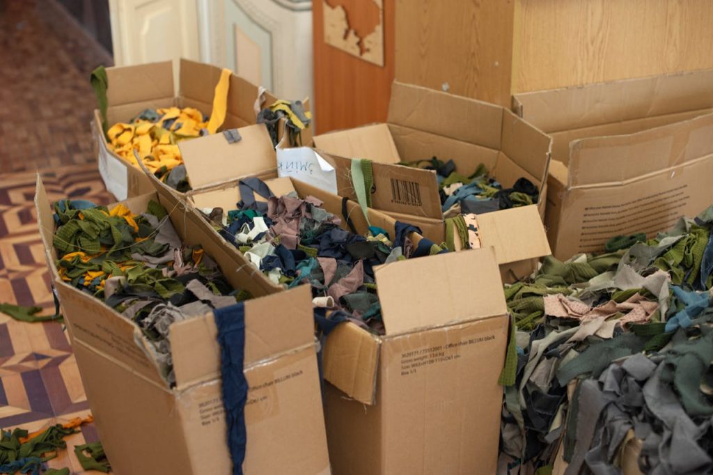 Cardboard boxes filled with multicolored fabric scraps in various textures and sizes, placed on a patterned wooden floor near a wall and furniture