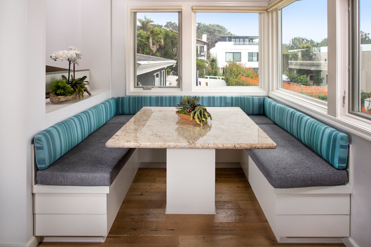 Bright breakfast nook with turquoise striped cushioned benches, granite table, and large surrounding windows overlooking a lush garden