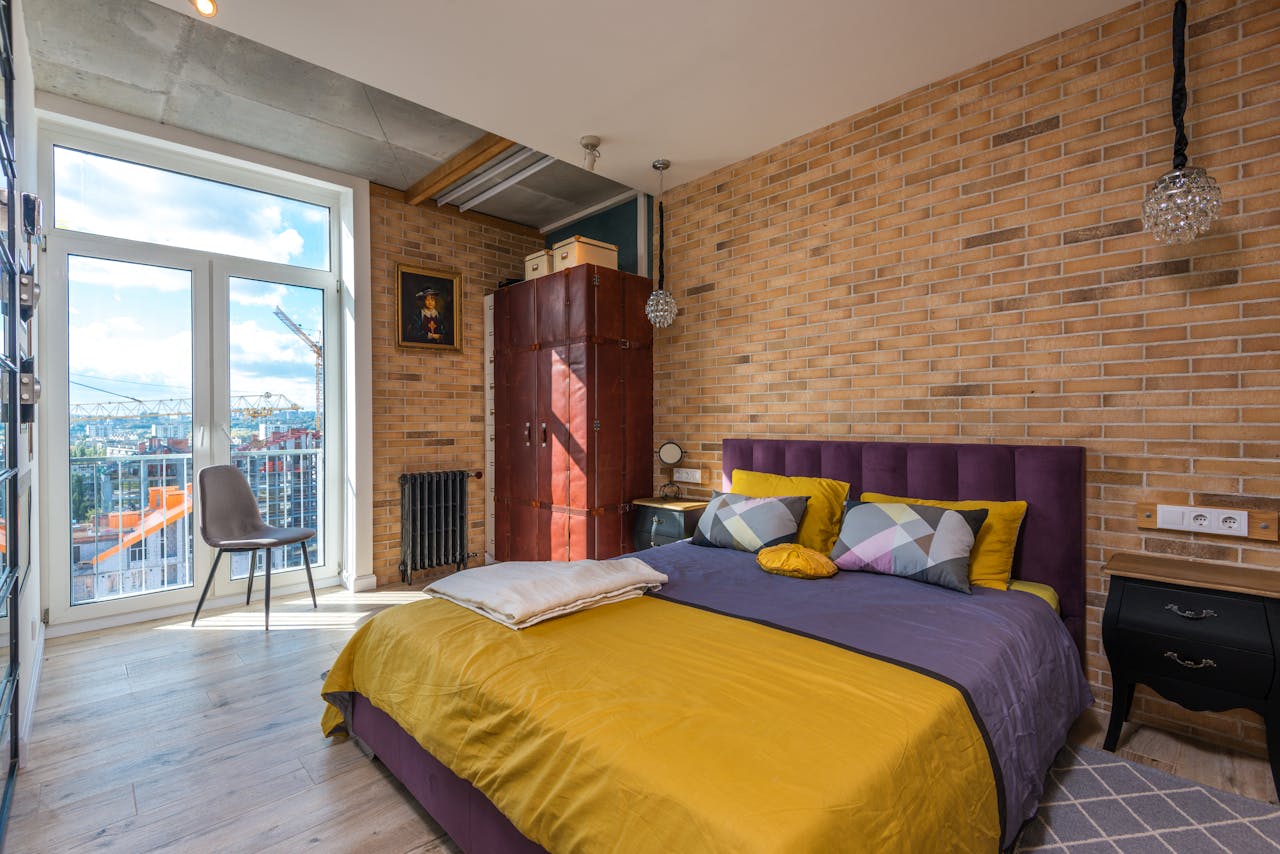 Modern bedroom interior, exposed brick wall, large window with city view, purple upholstered bed frame, mustard yellow and gray bedding, decorative pillows, ceiling-mounted pendant lights