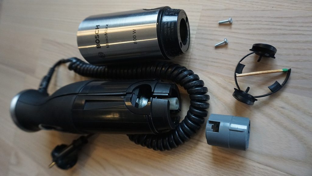 Disassembled Bosch MaxoMixx 800W hand blender, black and silver casing, exposed internal components, coiled power cord, two screws, black plastic retaining ring, gray connector part, wooden matchstick