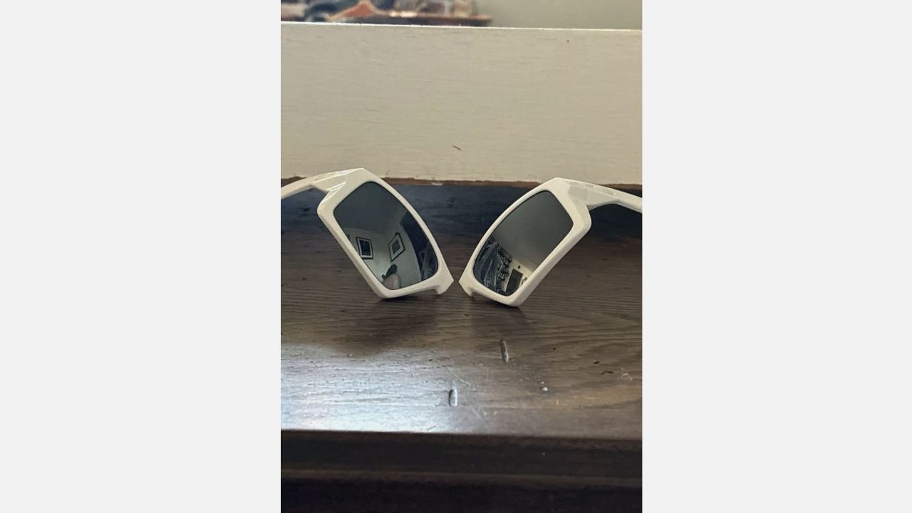 White sunglasses broken in half at the nose bridge, both lenses facing upward, reflecting a room interior, placed on a wooden surface