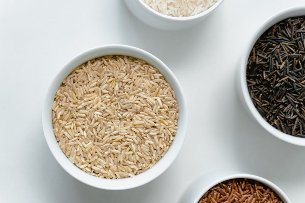 Close-up of assorted grains and seeds in white ceramic bowls, arranged on a white surface, including light brown, black, and reddish varieties, showcasing texture