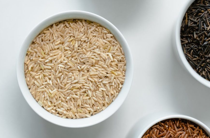 Close-up of assorted grains and seeds in white ceramic bowls, arranged on a white surface, including light brown, black, and reddish varieties, showcasing texture