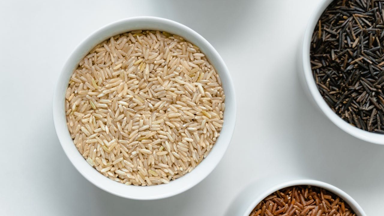 Close-up of assorted grains and seeds in white ceramic bowls, arranged on a white surface, including light brown, black, and reddish varieties, showcasing texture