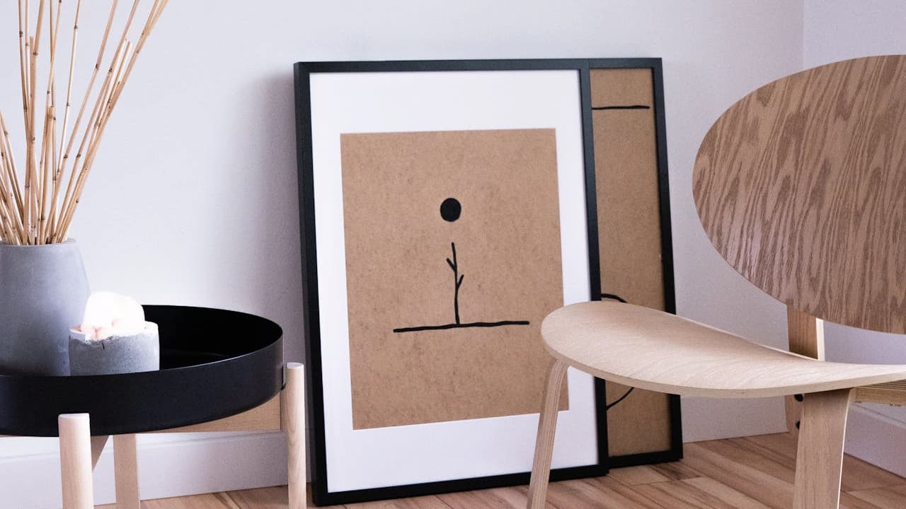 Minimalist interior setup, framed abstract artwork leaning against a white wall, modern wooden chair with woven backrest, black side table with a beige vase and dried grass, light wooden floor, clean and simple Scandinavian-style décor.