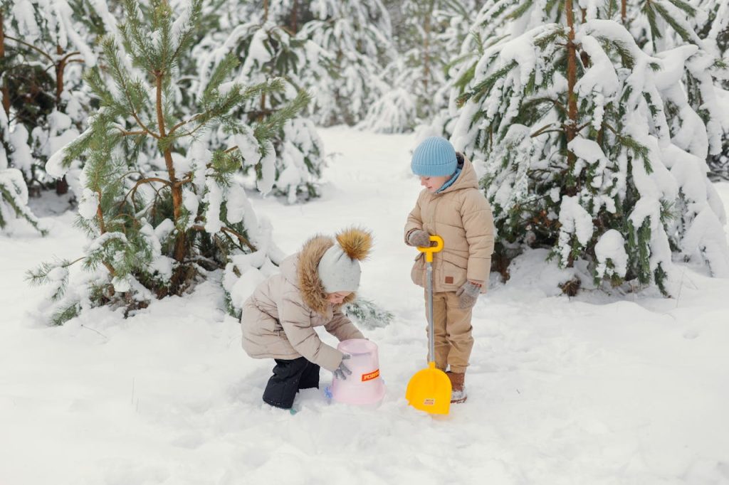 Two children bundled in winter clothing playing in the snow, building with a pink mold and yellow shovel among snow-covered trees