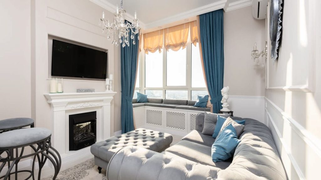 Elegant living room with tufted gray sectional sofa, blue and white accent pillows, wall-mounted TV above a white fireplace, chandelier lighting, large window with orange valance and blue curtains