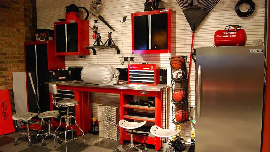 A well-organized garage workspace featuring red and black built-in Craftsman cabinets, a stainless steel workbench with tools and a toolbox, several industrial stools, a vertical sports ball rack, and a silver refrigerator