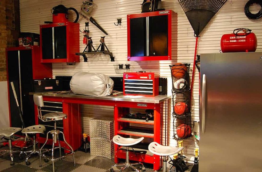 A well-organized garage workspace featuring red and black built-in Craftsman cabinets, a stainless steel workbench with tools and a toolbox, several industrial stools, a vertical sports ball rack, and a silver refrigerator