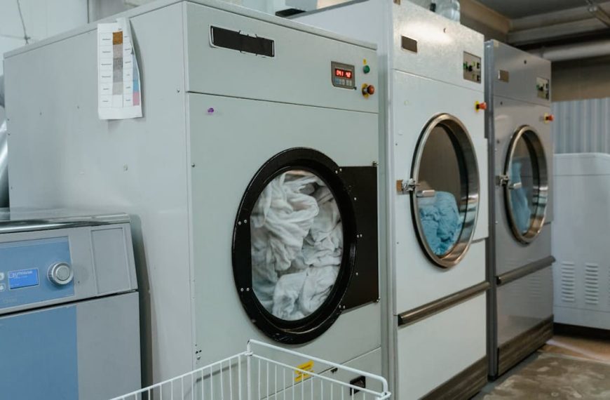 A row of large industrial washing machines in a laundry facility, with one machine filled with white linens and another with blue fabrics, indicating a bulky wash in progress