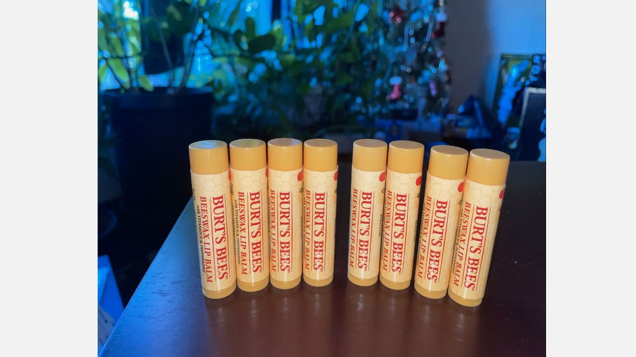 Nine tubes of Burt’s Bees Beeswax Lip Balm standing upright on a dark brown surface