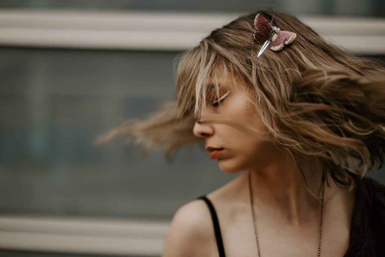 Young woman with blonde hair in motion, wearing a butterfly-shaped hair clip, black strap top, and necklace, standing against a blurred glass background