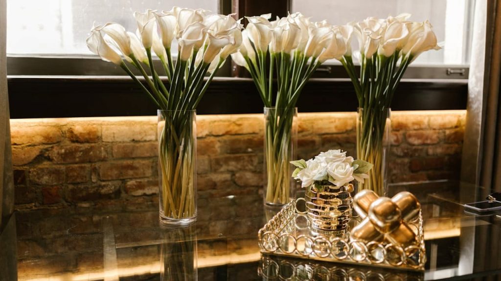 White calla lilies in tall glass vases, arranged on a glass table, in front of a window with natural light, brick wall background, decorative gold tray with white roses and metallic ornaments, elegant and modern interior décor