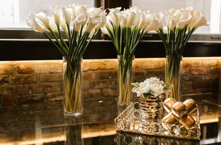 White calla lilies in tall glass vases, arranged on a glass table, in front of a window with natural light, brick wall background, decorative gold tray with white roses and metallic ornaments, elegant and modern interior décor