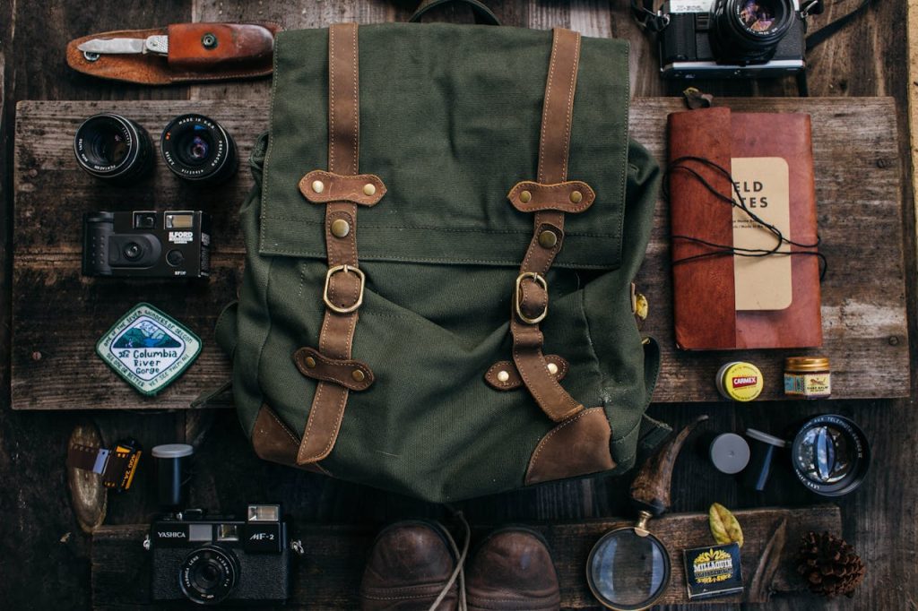 Green canvas backpack, surrounded by vintage cameras, lenses, travel journal labeled “Wild Notes,” compass, leather knife, boots, and small tins, all arranged neatly on a rustic wooden surface