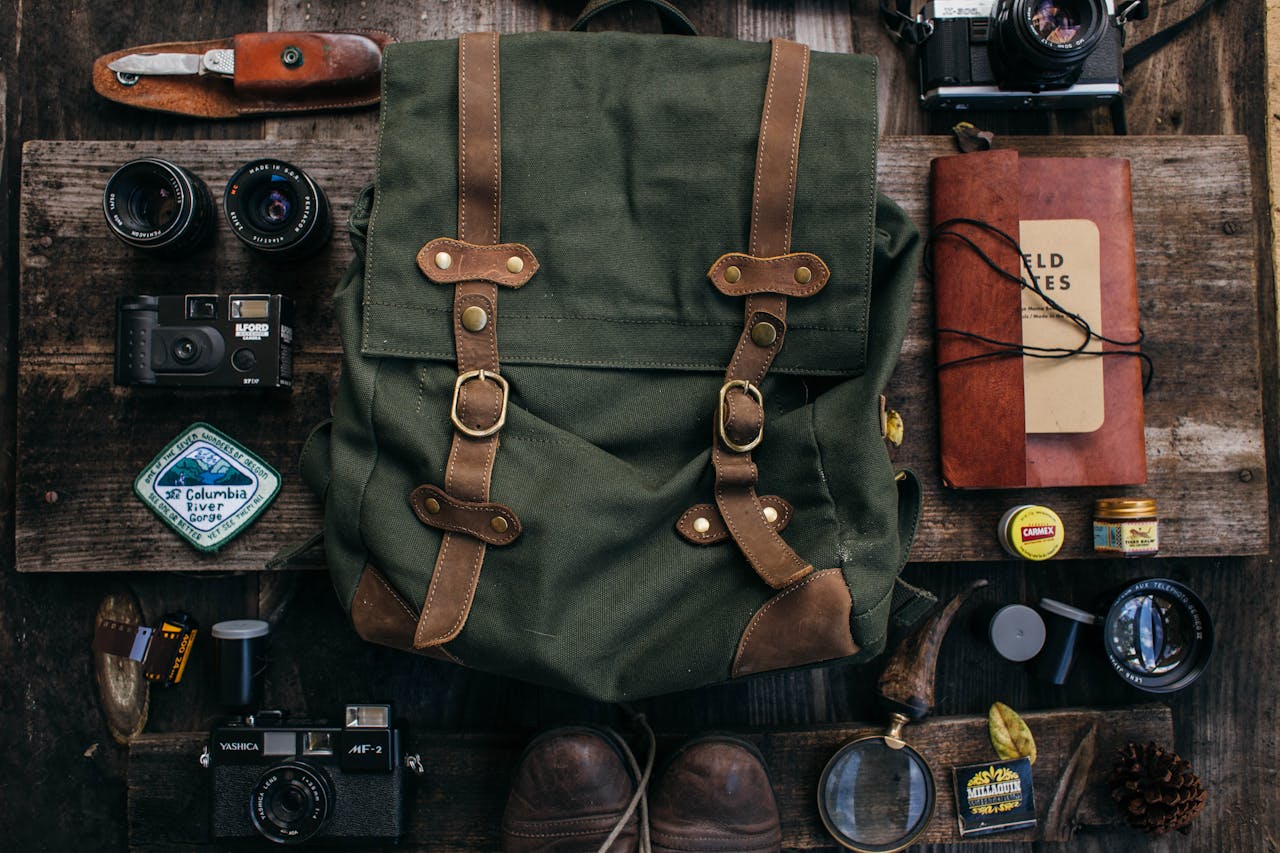 Green canvas backpack, surrounded by vintage cameras, lenses, travel journal labeled “Wild Notes,” compass, leather knife, boots, and small tins, all arranged neatly on a rustic wooden surface