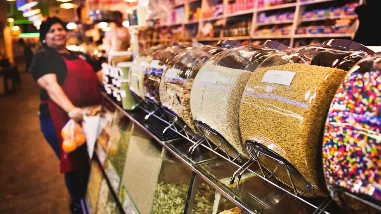 Colorful bulk bins filled with grains, seeds, and rainbow sprinkles line a counter in a store, with a smiling woman in a red apron holding produce in the background