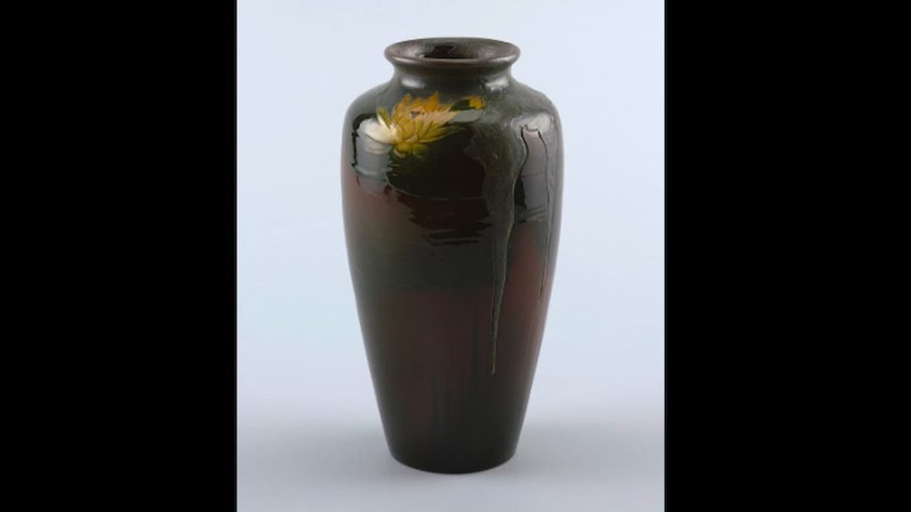 Tall ceramic vase with a glossy dark glaze, subtle green drips cascading from the neck, and a painted yellow water lily near the top, placed against a plain light background