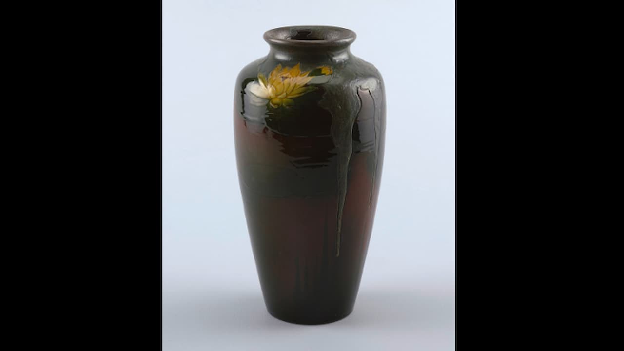 Tall ceramic vase with a glossy dark glaze, subtle green drips cascading from the neck, and a painted yellow water lily near the top, placed against a plain light background