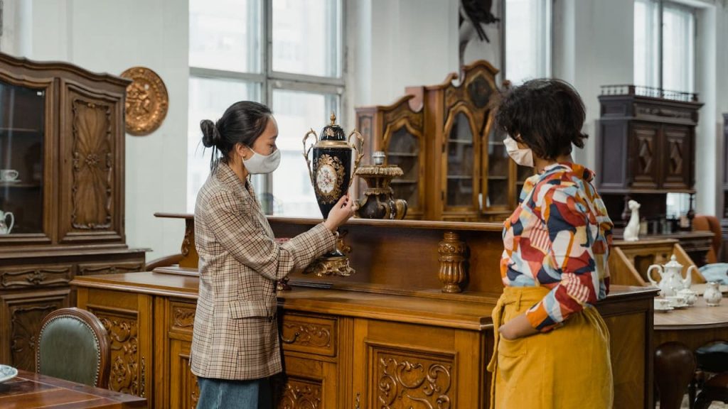 Two women wearing face masks, examining a decorative vase in an antique store, surrounded by ornately carved wooden furniture, vintage cabinets, and classic decor