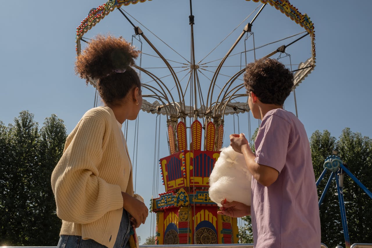 Two kids at a carnival standing in front of a colorful swing ride, one holding cotton candy