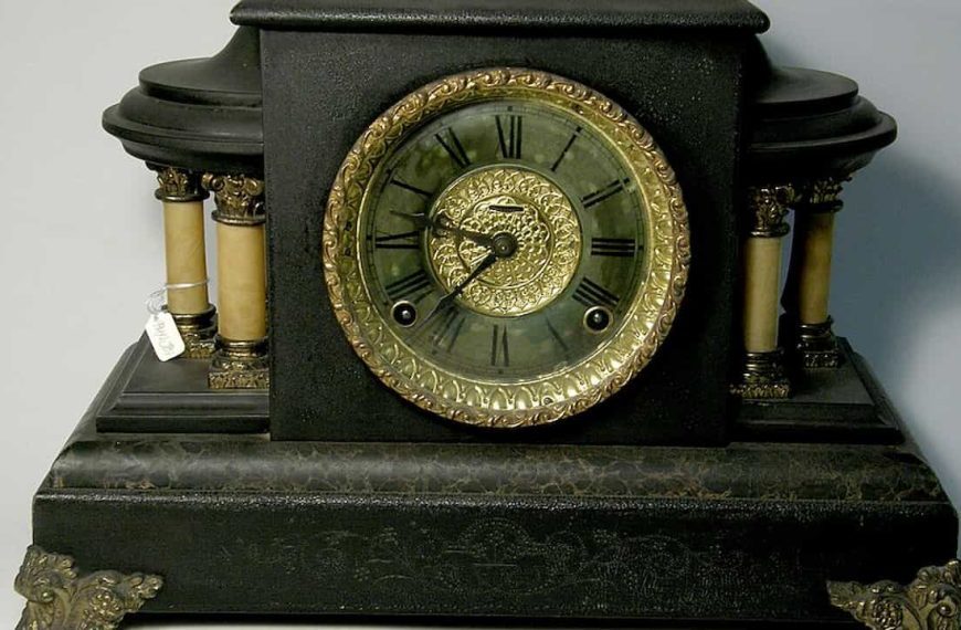 Black cast iron mantel clock with ornate gold-trimmed round face, Roman numerals, ivory side columns, decorative brass feet, intricate central dial pattern