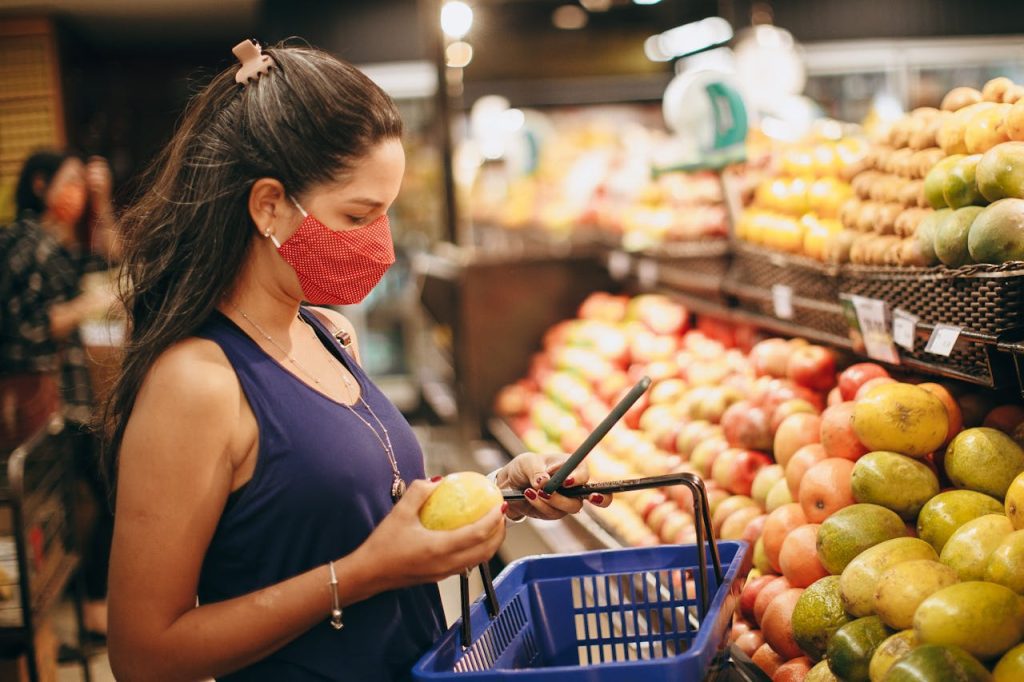 Woman wearing a red face mask, holding a mango in one hand and a phone in the other, standing in front of a fruit display in a grocery store, blue shopping basket hanging from her arm, checking product details or prices, surrounded by apples and mangoes