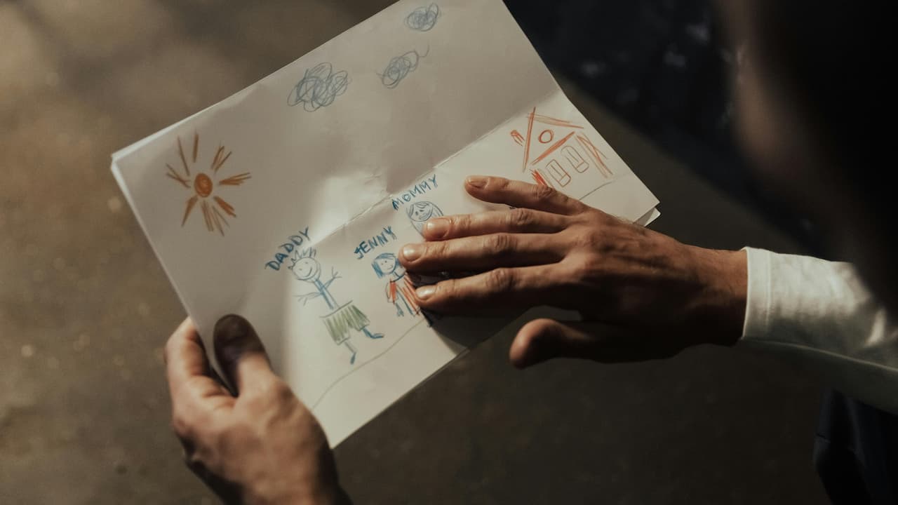 Person holding a child’s drawing showing stick figures labeled Daddy, Jenny, and Mommy, with a house, sun, and clouds drawn on the page