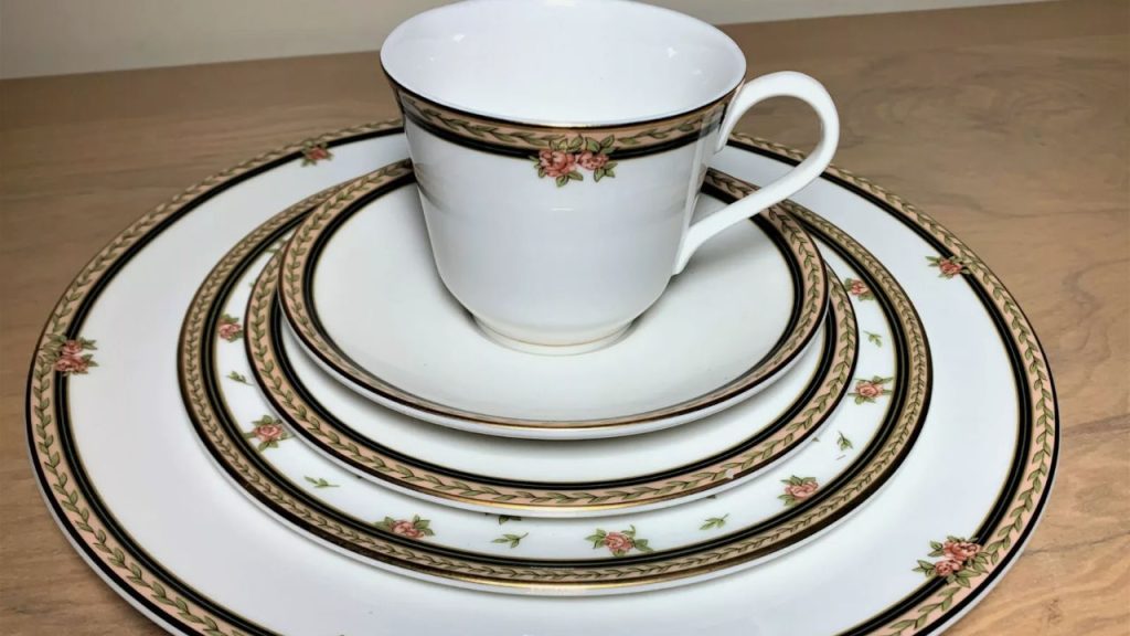 Elegant white china set with pink rose and green leaf border, including a teacup, saucer, and dinner plates, arranged on a wooden surface