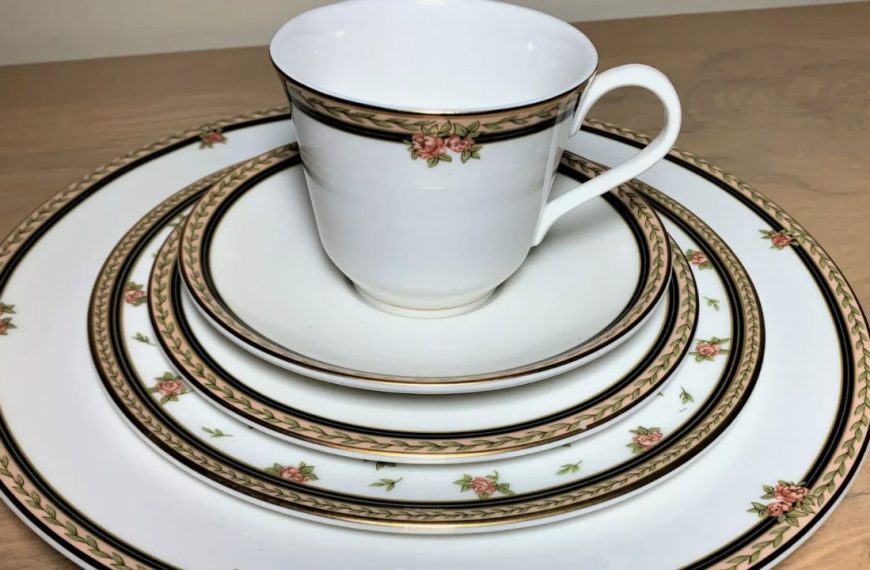 Elegant white china set with pink rose and green leaf border, including a teacup, saucer, and dinner plates, arranged on a wooden surface