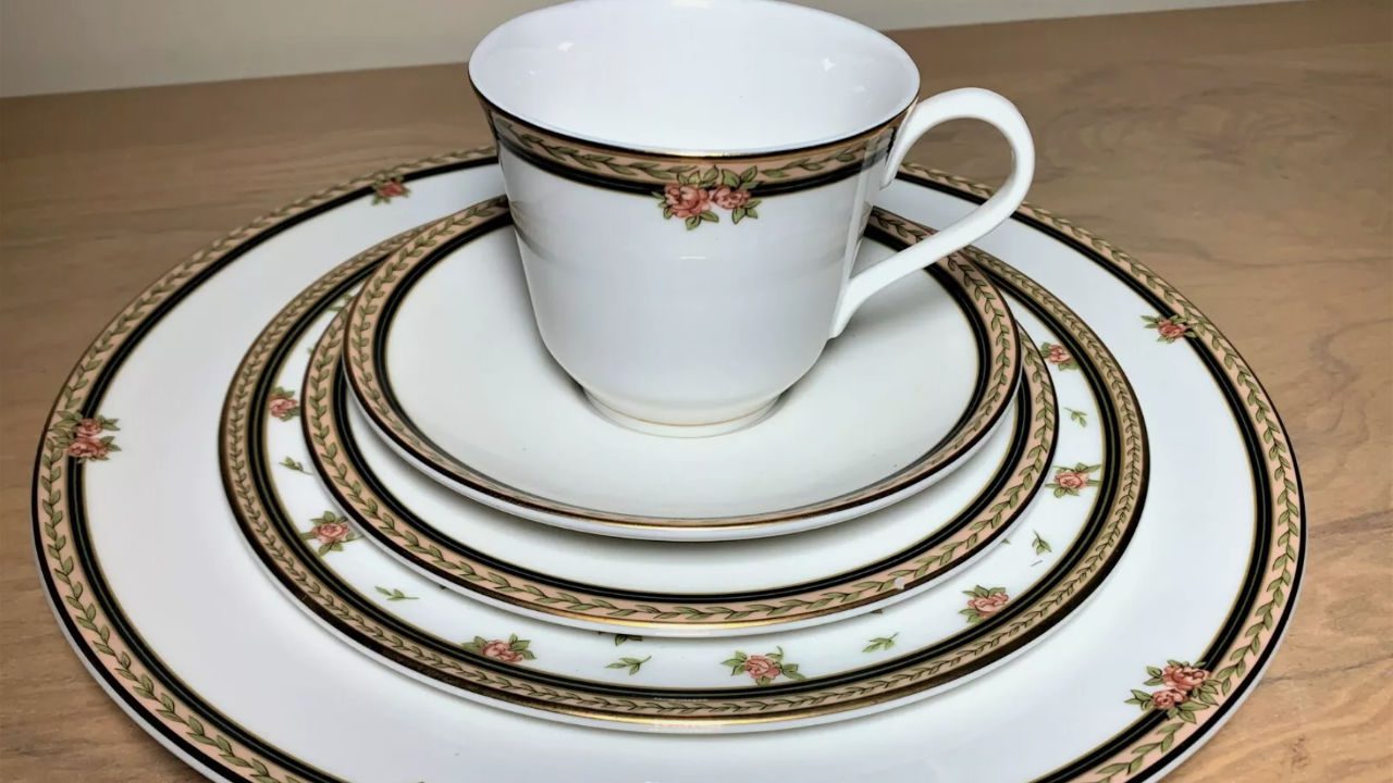 Elegant white china set with pink rose and green leaf border, including a teacup, saucer, and dinner plates, arranged on a wooden surface