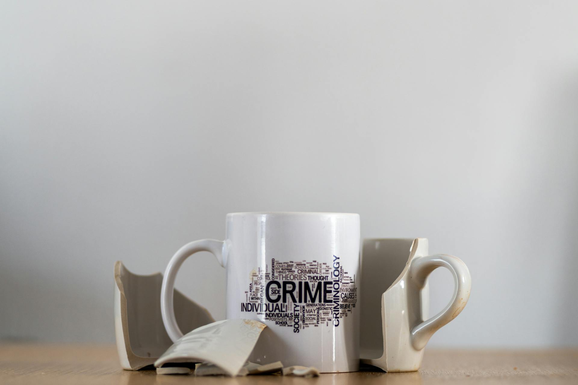 hree broken beige mugs surrounding a single intact white mug with a printed design, placed on a wooden surface, against a plain light gray background