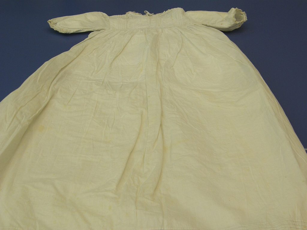 Vintage fabric dress, displayed on a dark blue background, featuring a gathered waist and tied bow at the top, textured material with subtle floral patterns, likely historical or ceremonial attire.
